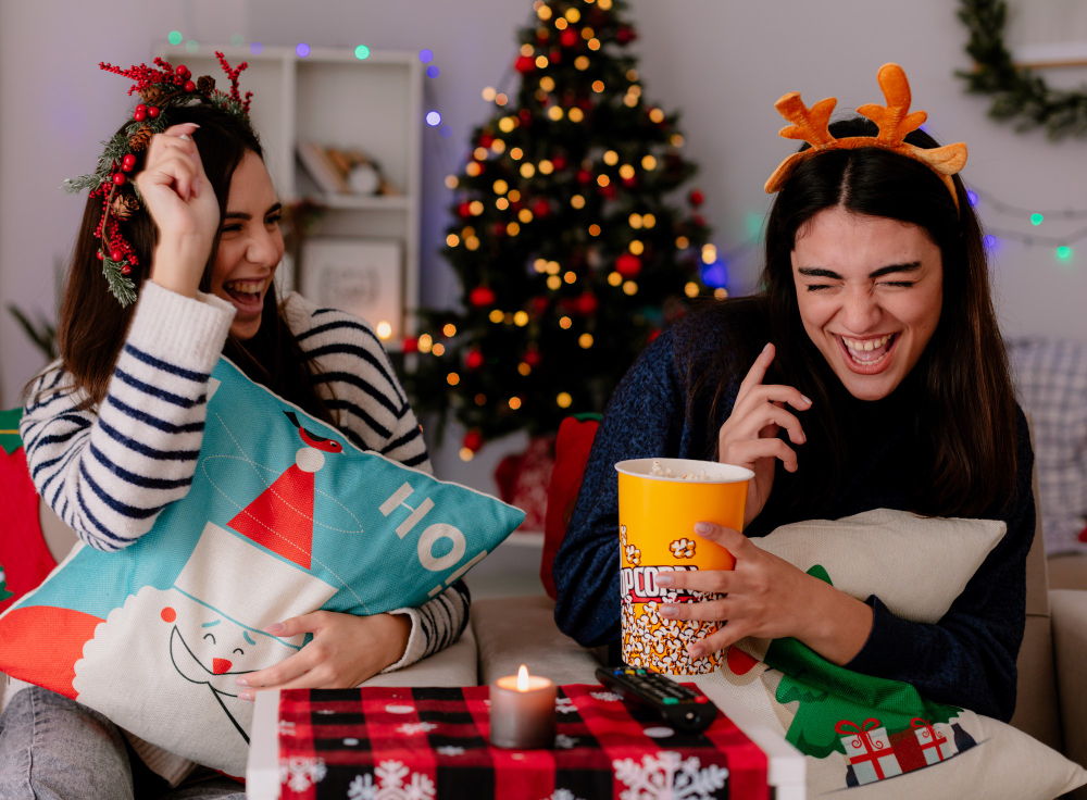 joyful pretty young girls with holly wreath and reindeer headband eat popcorn and play sitting on armchairs