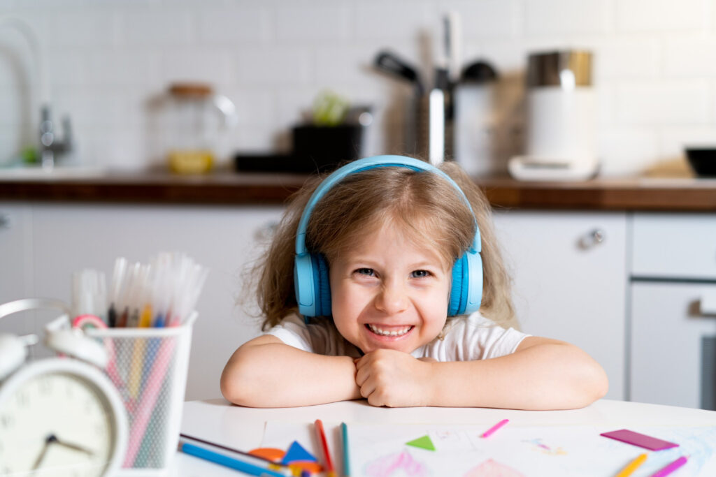 Little blonde girl at home in the kitchen at the table with headphones, draws and learns remotely.